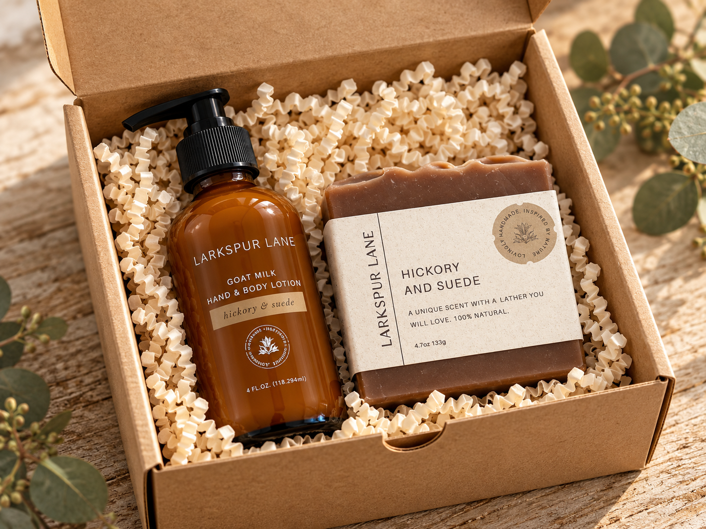 A gift set from Larkspur Lane in a brown kraft box filled with white packing paper. The set includes a bottle of Goat Milk Hand & Body Lotion and a bar of Hickory and Suede soap, styled with eucalyptus leaves on a wooden surface.
